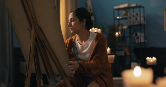 A beautiful woman dressed in curved clothes with hair pinned up sits on a chair at an easel with a canvas at home, pondering how to make a painting, a drawing, making precise movements