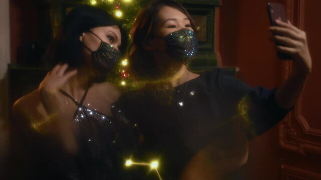 Two young women in shiny face masks take selfies on smartphone near decorated Christmas tree. Female couple smiles and takes pictures at a New Year's eve party at home