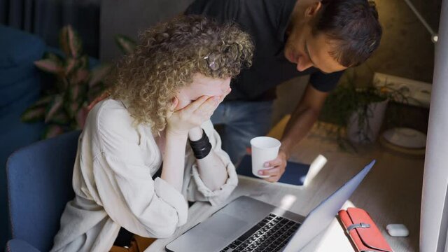 Tired woman cries sitting at modern laptop and careful husband gives cup of drink to calm lady down at work on hard project in home office closeup