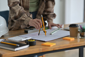 Cropped image of architect woman is drawing on a construction blueprint with a drawing compass and ruler.