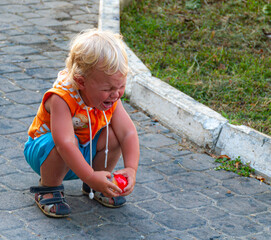 A small blond curly-haired boy in shorts and an orange blouse is squatting and crying loudly from...