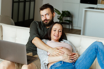 Pregnant wife in pink top and jeans lies on white sofa near sitting husband holding laptop on knees and smile looking into gadget