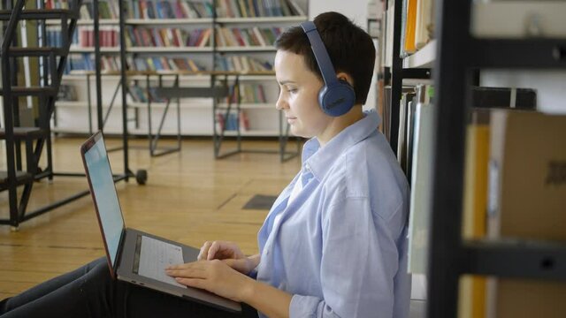 Short haired lady in blue shirt with headphones works on modern laptop to prepare for university tests in spacious light public library side view