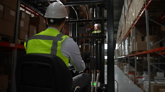 Rear view of forklift truck driver wearing hard hat and vest riding between racks filled with pallets of cardboard boxes. Bearded operator of loader during work in factory storage