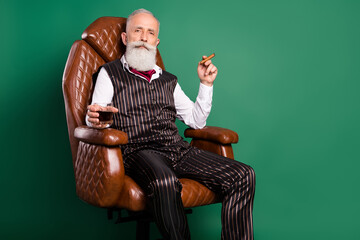 Photo of old noble aristocrat sit armchair drink whiskey enjoy cigar wear striped suit on green...