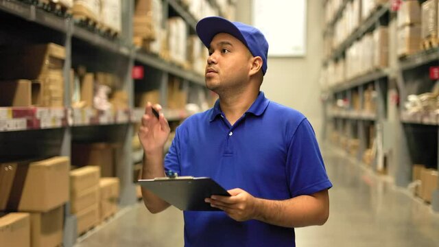 Distribution warehouse. Young worker in blue uniform checklist manage parcel box product in warehouse. Asian man employee holding clipboard working at store industry. Supply chain import export