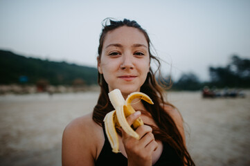 Portrait of a brunette girl eating a banana on a sandy beach in front of a hill