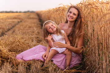 Naklejka na ściany i meble Mother and little daughter have fun sitting by the golden wheat field. Young woman tickles the girl
