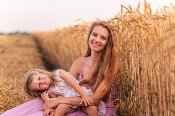 Mother and little daughter have fun sitting by the golden wheat field. Young woman tickles the girl