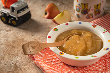 Applesauce in a plate with a child's drawing and a wooden spoon on a textured napkin with a mug and a toy car. Meals for children prepared at home..