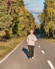 Senior active woman with gray hair running along forest paved road on sunny autumn day.