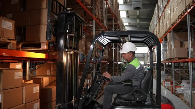 Close-up of warehouse worker operating forklift, placing pallet full of cardboard boxes on shelf. Professional forklift driver lifting goods on rack filled with cardboard boxes