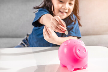 Happy and smiling Asian little toddler girl putting coin into Piggy Bank for personal saving the money is Sitting On Sofa At Home in the morning. Studio shot Copy Space. Child money-saving concept