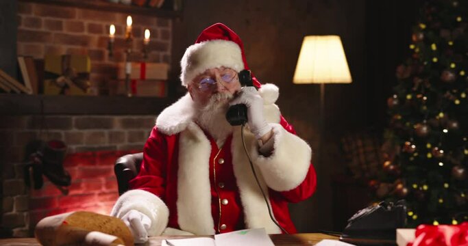 Santa Claus working at night, calling on old phone close-up, lots of letters from kids. St Nicholas preparing presents for kids. Late work on Christmas Eve. Green tree and fireplace on background.
