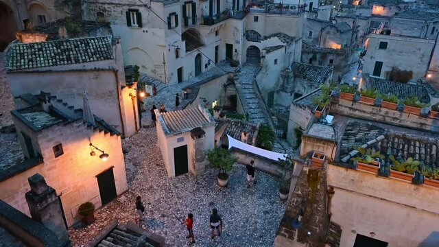 Matera, Basilicata, Italy. August 2021. Footage at blue hour in the stones, the historic center of the village. A few passers-by in the alleys illuminated by yellowish light. Tilt over the cityscape.