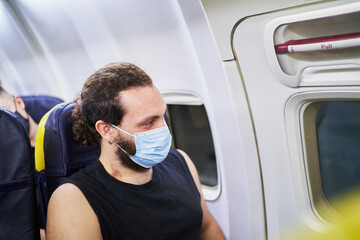 Fototapeta na wymiar Cheerful adult man with surgical mask looking through the window of a plane