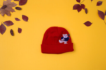 Fashionable children's clothing. Cute baby red hat with bear on yellow background with leaves. Outfit for a little boy. Winter, autumn collection. Organic cotton. Top view, flat lay, autumn baby look