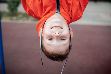 A boy in autumn clothes hangs upside down on the playground.