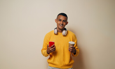 young latinx man with headsets holding with the hands a mobile phone and coffee cup, winter concept.
