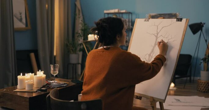 A woman with long brown hair pinned up in a bun using pencils sits by candles in the evening. A focused girl with the soul of an artist draws a picture of a tree, sketches on a cavas