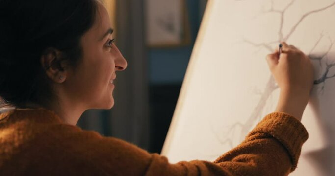 An art student sits in her home living room in the evening and draws a sketch of a tree, nature for a college credit. The passionate woman smiles, satisfied with painting