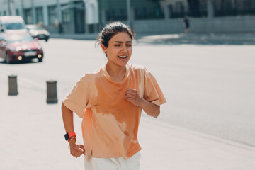Young indian woman runner jogging in wet sweaty t-shirt at city street urban