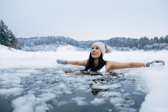 Winter cold swimming. Woman in frozen lake ice hole. Swimmers wellness in icy water. How to swim in cold water. Beautiful young female in zen meditation. Hat and gloves swimming clothes. Nature lake