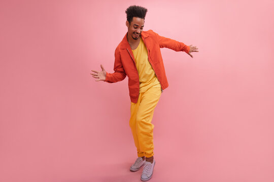African teenager guy actively moves to beat of music on pink background. In bright outfit from mustard T-shirt, vintage jeans with orange jacket, posing. Concept of enjoying moments. - Powered by Adobe