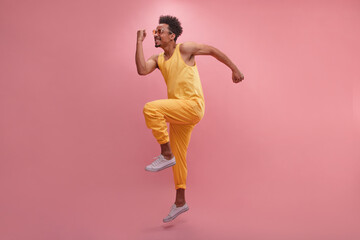 Fototapeta na wymiar Swarthy young African guy half-sideways jumping with happiness in place with copy space. Male model wearing sunglasses wears yellow tank top and pants. People sincere emotions lifestyle concept.