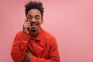 Obraz premium Close-up portrait positive dark-skinned young guy closing eyes and recalling something on pink background. Brunette with afro hair gently touches face and smiles, wearing jumpsuit and glasses.
