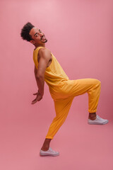 Fancy dark-skinned young man in yellow suit lifted legs and rejoices with copy space. Cute boy looking at camera and ready to have fun posing in front pink wall. Emotions people and fashion concept.