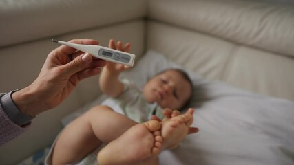 Worried young mother sitting on sofa beside her sick son with high fever. Mom measures temperature using thermometer of sick child lying under blanket at home.