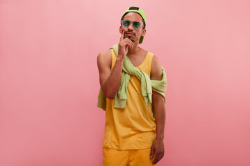 Student against pink wall express pensive emotion, touching chin, looks aside. Medium shot of thoughtful impressive young brown-skinned man with green eyewear, dressed up colourful. 
