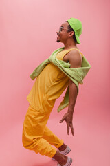 Energetic young, dark-skinned man stands half-turned and tries to touch legs from behind. Posing against crimson background in bright yellow outfit with green long sleeve on shoulders and cap.