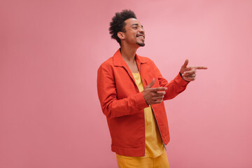 Handsome African young man with curly dark hair points his index fingers to side. Dressed in red jacket, yellow jeans and T-shirt, laughs while talking at someone's joke. Concept spread good mood