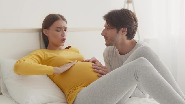 Worried husband comforting his pregnant wife, woman suffering from acute pain, lying on bed at home