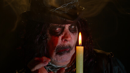 Fototapeta premium Frightening senior woman with Halloween witch makeup looking at candle, conjure, hex, wiz. Horror theme. Sinister sorcerer lady on studio black background. Voodoo magical creepy rituals with candle