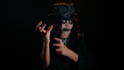 Frightening senior woman with Halloween witch makeup listening music, smiling dancing to disco music rhythmically moving her hands, having fun. Sinister lady on studio black background. Voodoo rituals