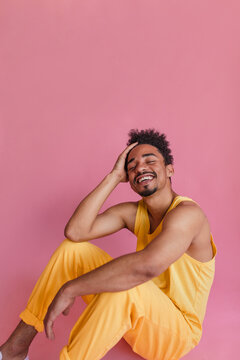 Full-length Shot Of Attractive Afro Man Sitting On Floor In Pink Room. Posing Naturally And Smiling Broadly With Her Snowy Teeth In Pastel Yellow Pants And Tank Top.