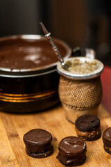 Homemade Argentine Alfajores dipped in chocolate on a wooden board next to a yerba mate tea and the pot containing the chocolate