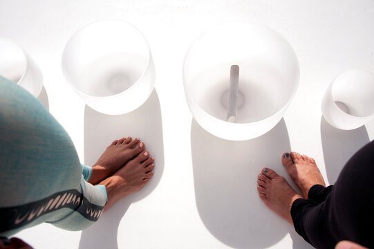 The Feet Of Two Women Standing Over Crystal Healing Bowls. Sound Therapy, Vibrational Medicine, Is A Tool Used To Help Promote Self-healing. 