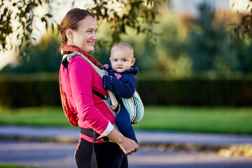Happy european woman with little boy in sling walks in park.