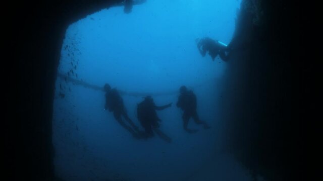 Silhouette of divers with torches entering a large chamber of a deep underwater shipwreck.