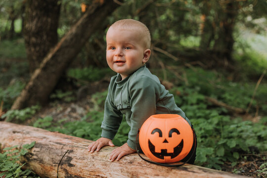 portrait of cute baby boy holding on to a log against the background of a green forest. pumpkin basket for sweets in the foreground. walking and playing outdoor. Halloween concept. High quality photo