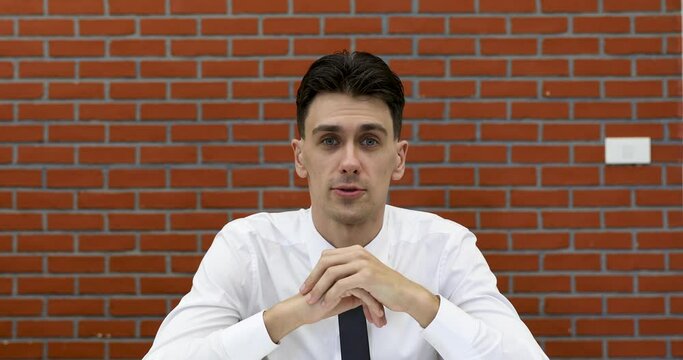 Young adult businessman in casual white shirt sitting in front of red brick wall, raise hand to camera for greeting to audience in online conference and start to speak.