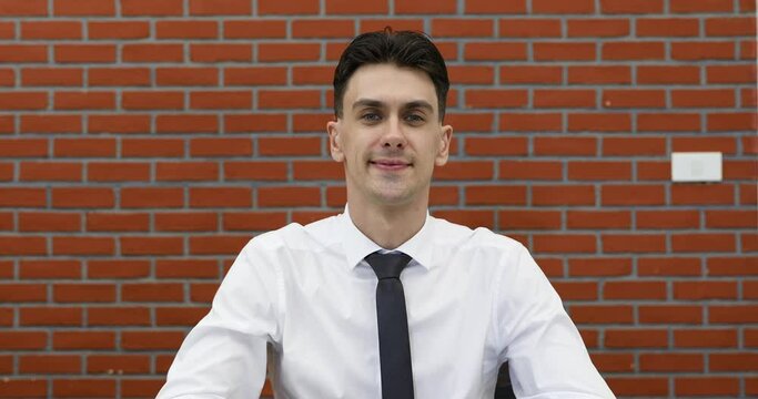 Young adult businessman in casual white shirt sitting in front of red brick wall, raise hand to camera for greeting to speaker in online conference and stay listening.