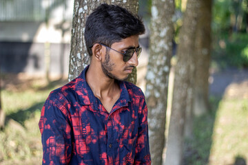 In the late afternoon sun, a boy peeks out from behind a tree and black sunglasses his eyes and...