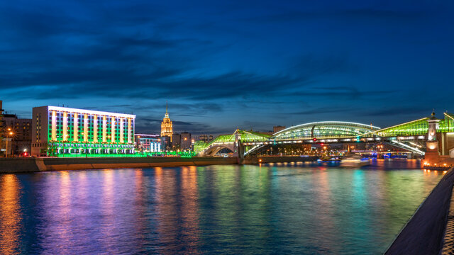 View of the colorful Bogdan Khmelnitsky bridge illuminated at night reflecting in the Moskova river. Moscow Kiyevsky railway station at night. Moscow, Russia