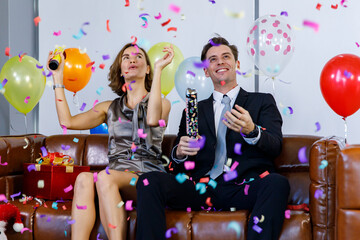 Caucasian businessman in business suit sitting on leather sofa with present gift box and balloons with female girlfriend in casual dress shoot paper confetti popper together celebrating xmas festival