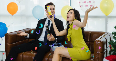 Caucasian male businessman in business suit and female businesswoman in formal dress sitting on leather sofa shooting colorful paper confetti popper together on Christmas eve festival office party